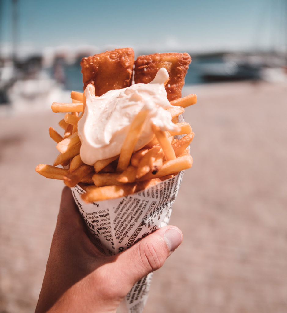 close up of a white person's hand holding a cone of fish and chips with a blurry beach in the background