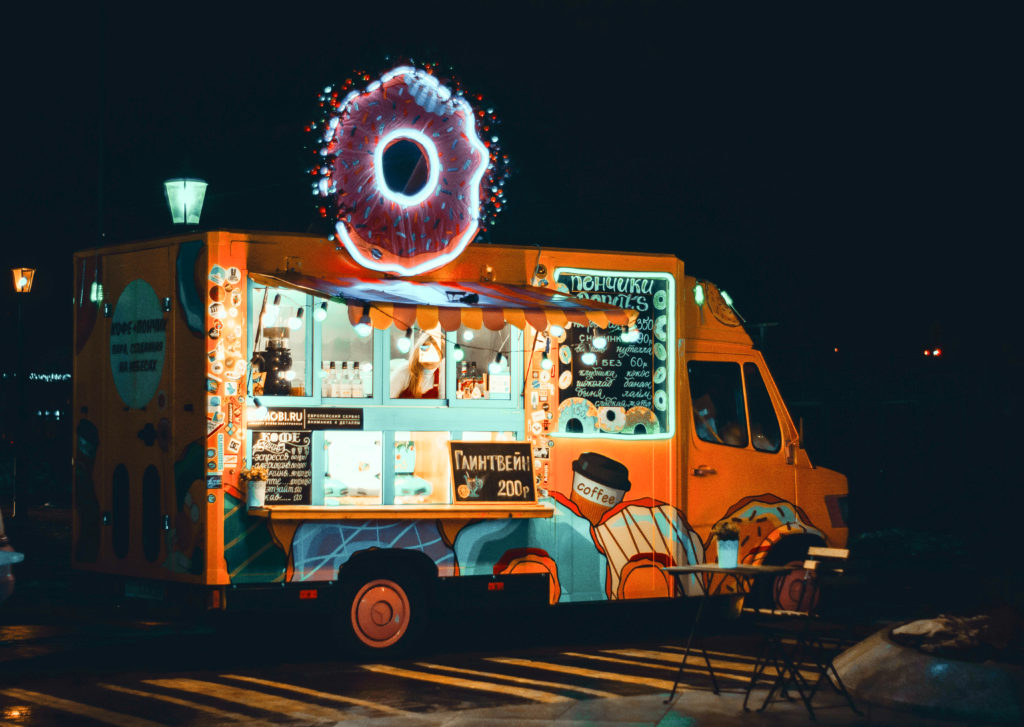 photo of an orange and blue food truck at night time with a neon donut sign